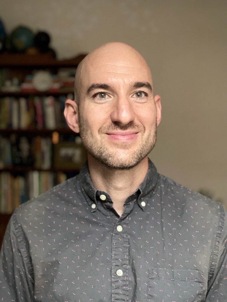 Man with short beard, wearing a grey button up shirt, standing in front of a bookshelf.
