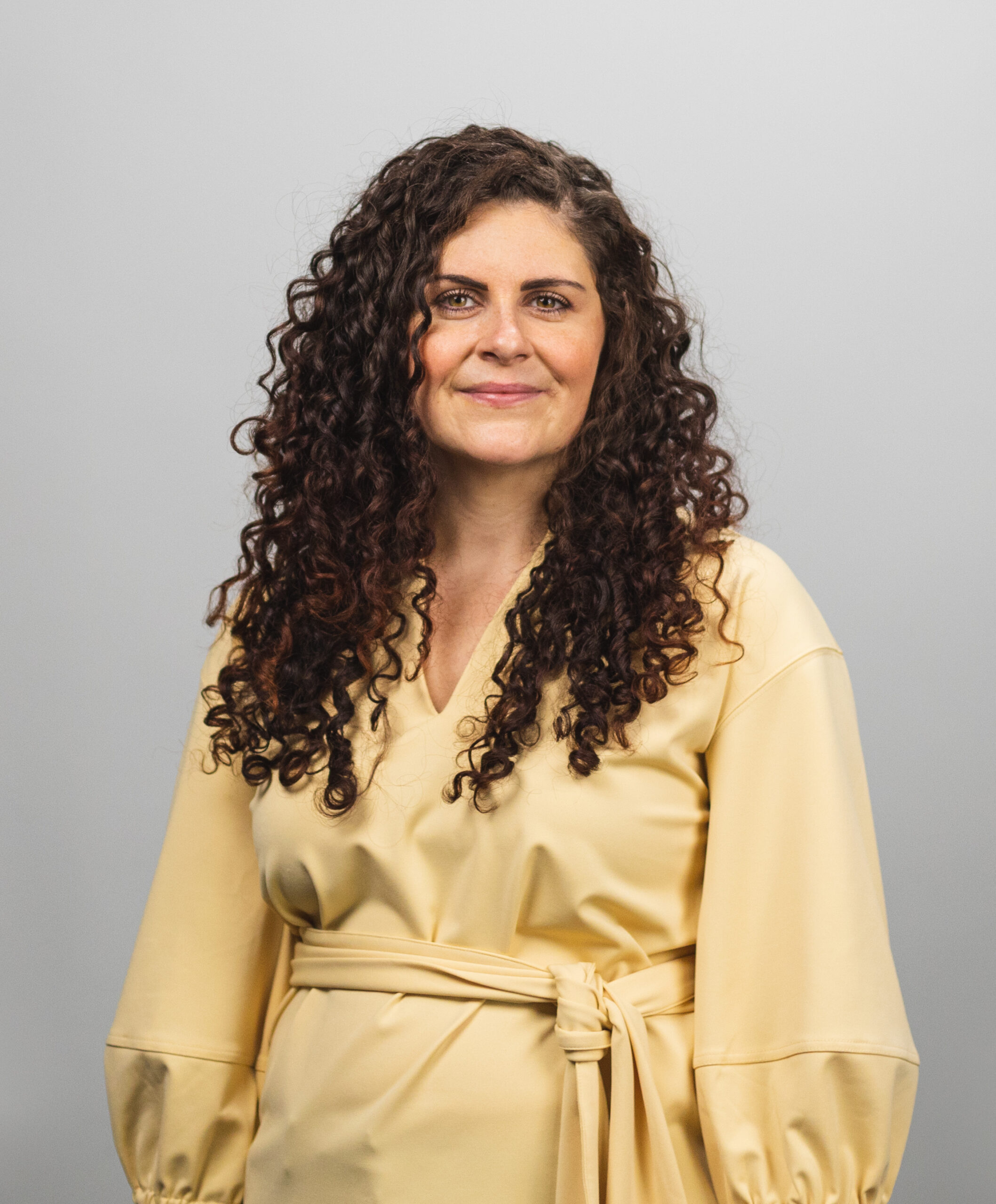 Woman with long curly brown hair wearing a pale yellow wrap around dress.