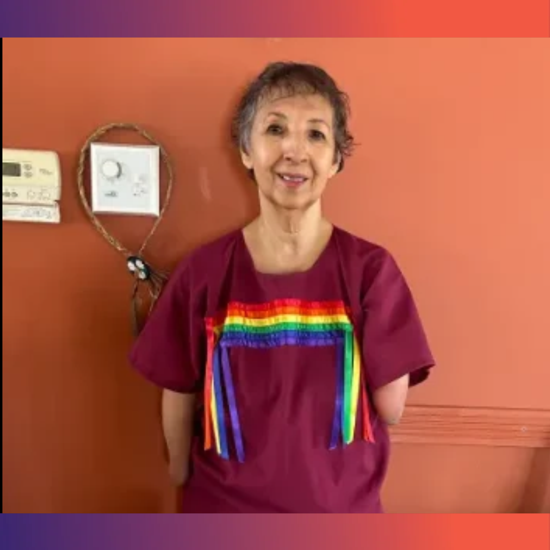 Photograph shows a smiling woman standing indoors against a coral wall, wearing a burgundy shirt with a bright rainbow design.